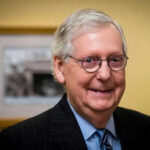Trump Allies DEMOLISH McConnell’s 40-Year Machine Smiling man in suit with round glasses indoors.
