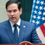A man in a blue suit speaking into a microphone with an American flag in the background