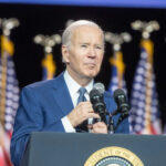 Man speaking at podium with flags behind him.