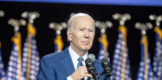 Man speaking at podium with flags behind him.