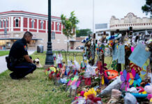Police officer kneels at a memorial with flowers and tributes.