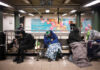 Individuals sitting on benches in a subway station, some covered with blankets