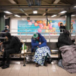 Individuals sitting on benches in a subway station, some covered with blankets