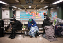 Individuals sitting on benches in a subway station, some covered with blankets