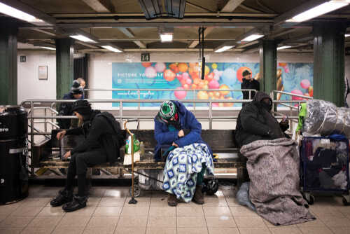 Individuals sitting on benches in a subway station, some covered with blankets