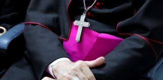 A close-up of a clergy member's hands clasped together, wearing a cross necklace and formal attire