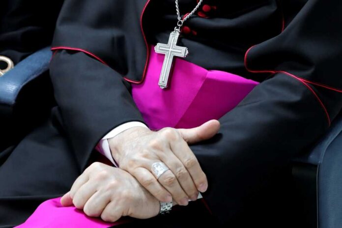 A close-up of a clergy member's hands clasped together, wearing a cross necklace and formal attire