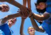 Group of female athletes joining hands in a circle under a blue sky