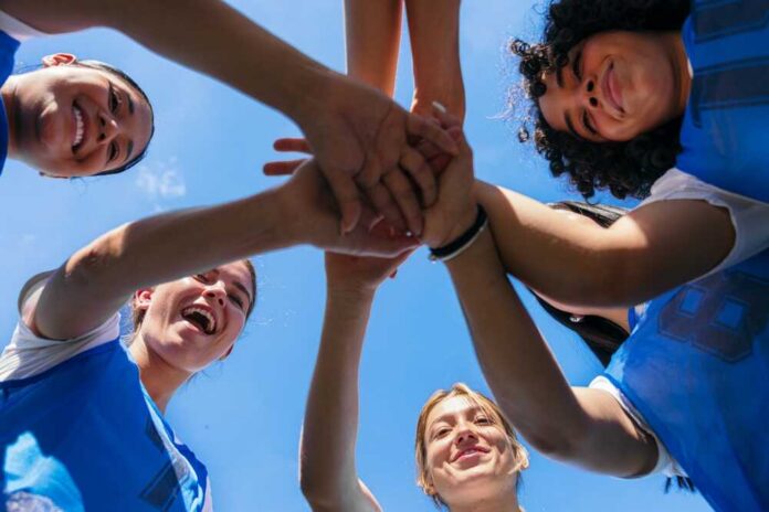 Group of female athletes joining hands in a circle under a blue sky