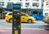 A parking meter on a city street with a yellow taxi passing by