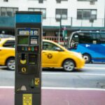 A parking meter on a city street with a yellow taxi passing by