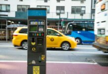 A parking meter on a city street with a yellow taxi passing by