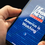Hand holding smartphone showing Fox News Channel logo.