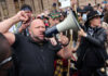 A man shouting into a megaphone during a protest with a crowd in the background