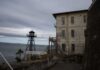 An old lighthouse next to a weathered building by the water under a cloudy sky