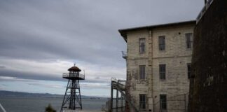 An old lighthouse next to a weathered building by the water under a cloudy sky