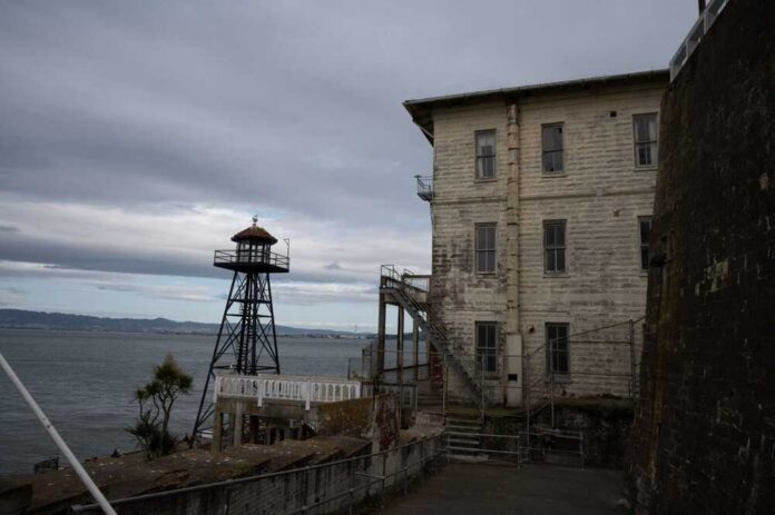 An old lighthouse next to a weathered building by the water under a cloudy sky
