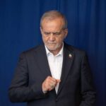 A man in a suit with a serious expression standing against a blue background