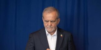A man in a suit with a serious expression standing against a blue background