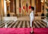 A woman in formal attire standing in a grand hall with flags in the background