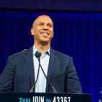 Party Panic: Booker Won’t Back Platner Man smiling at podium with American flag behind