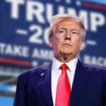 man in suit with red tie standing before a blurred campaign-style sign