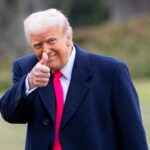 A man in a dark coat and pink tie giving a thumbs up outdoors