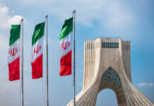 Drone Reports Over Tehran — Escalation Trap Brewing Three Iranian flags in front of the Azadi Tower against a blue sky