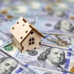 Tax The Rich Becomes Dox The Middle Mini wooden house on scattered US hundred-dollar bills