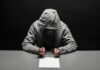 Person in gray hoodie writing while handcuffed at a table