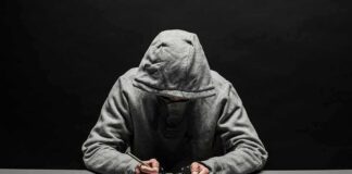 Person in gray hoodie writing while handcuffed at a table