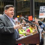 Pritzker’s ‘Easiest’ Claim Meets Tax Reality Speaker at a podium addressing a crowd with microphones and protest signs