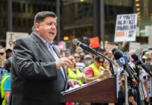 Speaker at a podium addressing a crowd with microphones and protest signs