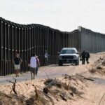 Border War Ignites: Madrid Hits Rome Back Border fence with patrol vehicles and people walking on a dirt road