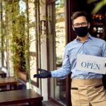 Masked waiter holding an open sign outside a café