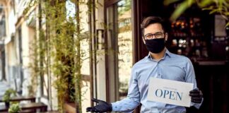 Masked waiter holding an open sign outside a café