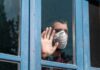 Autumn Covid Upswing: Calm Or Crisis? Person in a face mask looking out a window with hand raised