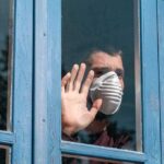 Autumn Covid Upswing: Calm Or Crisis? Person in a face mask looking out a window with hand raised