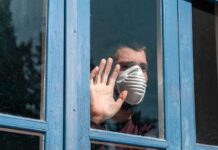 Autumn Covid Upswing: Calm Or Crisis? Person in a face mask looking out a window with hand raised