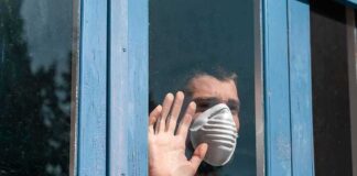 Person in a face mask looking out a window with hand raised
