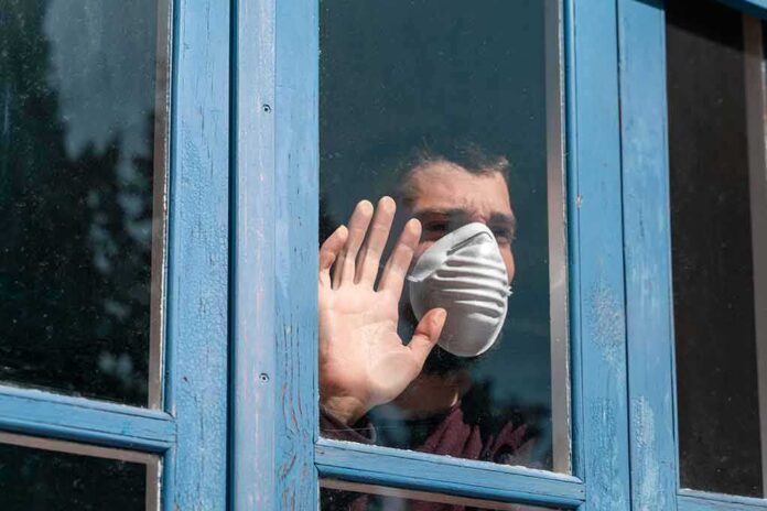 Person in a face mask looking out a window with hand raised