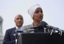 Brutal Regime Ties Shadow Omar – Did The Paperwork Hide This? Ilhan Omar speaking at a podium with André Carson standing behind